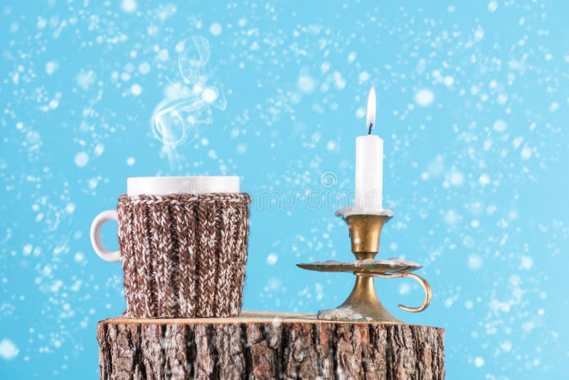 Retro cup of hot tea in wool scarf and candle in old candlestick burning on wood log. It`s snowing and blue background. Winter idyll and Christmas holiday concept. Retro cup of hot tea in wool scarf and candle in old candlestick burning on wood log. It`s snowing and blue background. Winter idyll and Christmas holiday concept