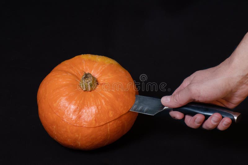 Hand holds a knife and cuts a round orange pumpkin, isolated on black background, close-up, copy space. Step-by-step instructions for preparing a jack lantern. Step 1. Hand holds a knife and cuts a round orange pumpkin, isolated on black background, close-up, copy space. Step-by-step instructions for preparing a jack lantern. Step 1