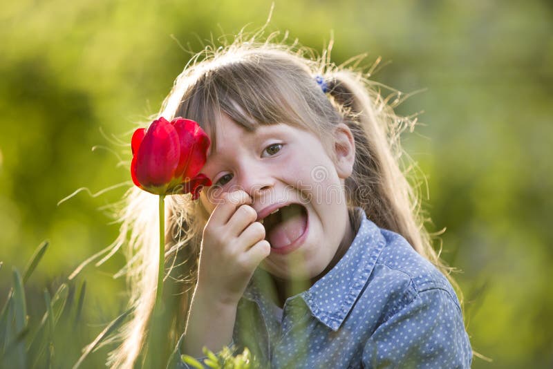 Cute pretty smiling child girl with gray eyes and long hair with bright red tulip flower on blurred sunny green bokeh background. Love to nature concept. Cute pretty smiling child girl with gray eyes and long hair with bright red tulip flower on blurred sunny green bokeh background. Love to nature concept.