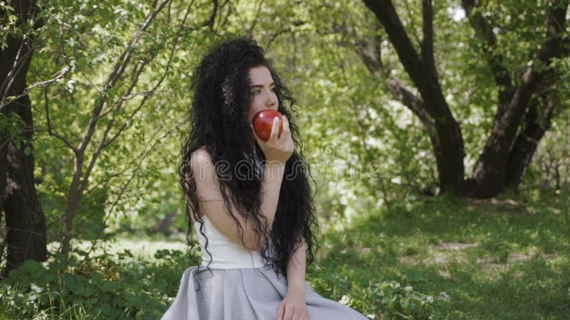 Το όμορφο brunette κάθεται στο θερινό κήπο και τρώει το κόκκινο μήλο