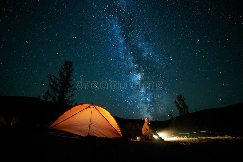 Tourist near his camp tent at night under a sky full of stars. Orange illuminated tent. Tourist near his camp tent at night under a sky full of stars. Orange illuminated tent.