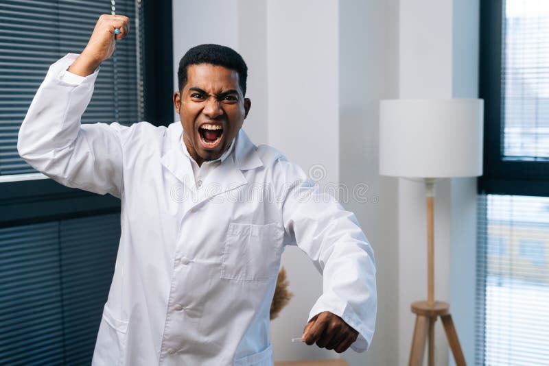 Mad African-American male doctor in white uniform screaming furious, shouting with anger holding in hand syringe, looking at camera. Concept of vaccination, immunization, disease prevention. Mad African-American male doctor in white uniform screaming furious, shouting with anger holding in hand syringe, looking at camera. Concept of vaccination, immunization, disease prevention.