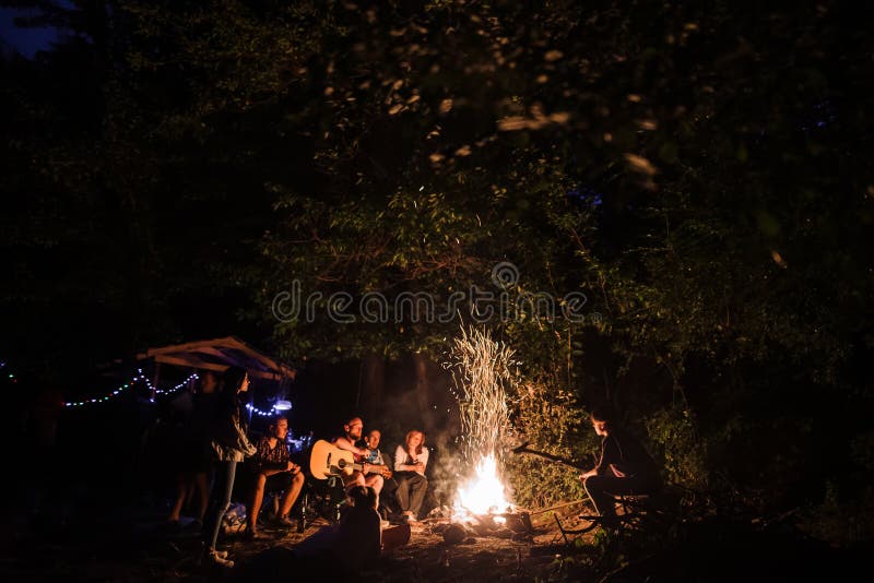 Φίλοι ταξιδιώτες που αράζουν στη μεγάλη φωτιά, τραγουδούν τραγούδια και παίζουν κιθάρα στην κατασκήνωση στο νυχτερινό δάσος Ομάδα στοκ εικόνες με δικαίωμα ελεύθερης χρήσης
