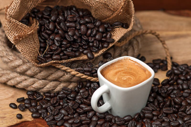 Espresso in white square espresso cup surrounded by coffee beans spilling over from a burlap bag. Espresso in white square espresso cup surrounded by coffee beans spilling over from a burlap bag