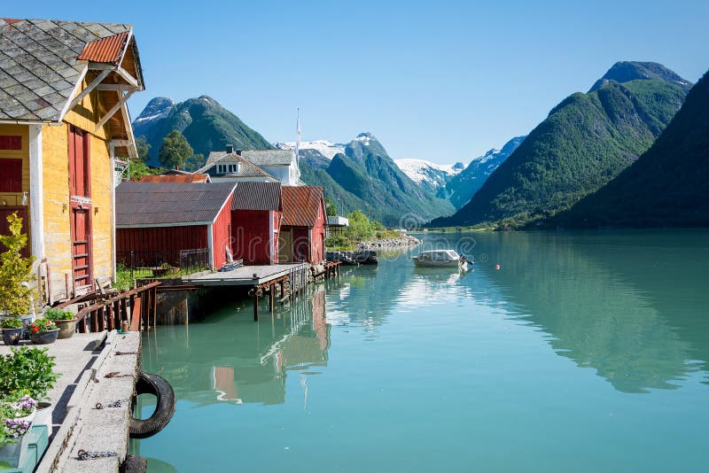 View over the fjord Fjaerlandsfjord and the small village of Mundal (or Fjaerland) with some snow-capped mountains, a boat, reflection in the water and yellow and red boathouses in Sogn of Fjordane, Norway. View over the fjord Fjaerlandsfjord and the small village of Mundal (or Fjaerland) with some snow-capped mountains, a boat, reflection in the water and yellow and red boathouses in Sogn of Fjordane, Norway.