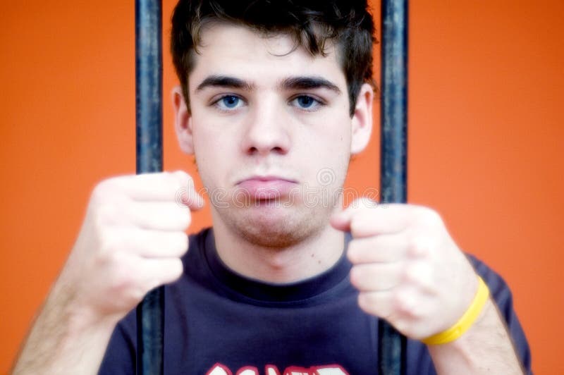 Young male behind bars. Sad faced grabbing bars in both hands. Looking forward. Orange background. Young male behind bars. Sad faced grabbing bars in both hands. Looking forward. Orange background.