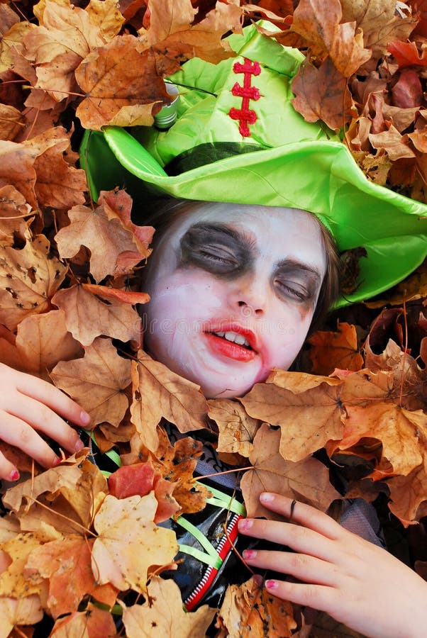 Young girl in Halloween makeup and costume plays dead in a pile of leaves; only her face and hands are exposed. Young girl in Halloween makeup and costume plays dead in a pile of leaves; only her face and hands are exposed.