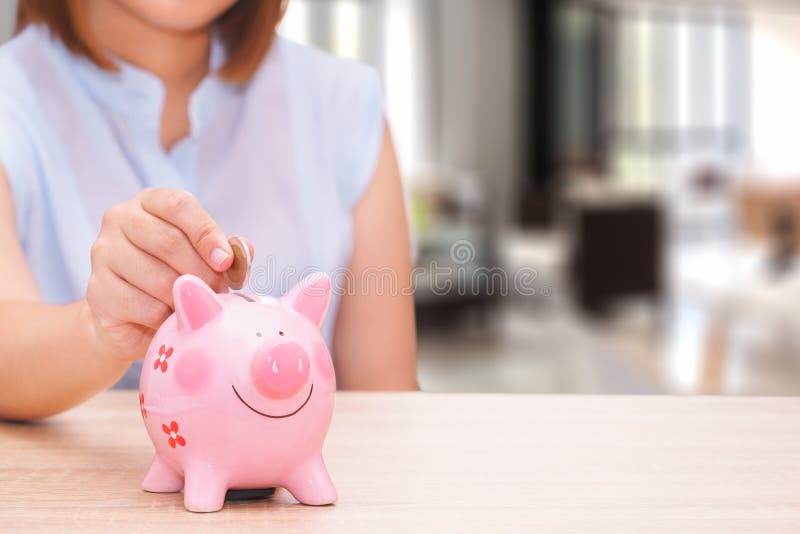 Χέρι γυναικών που βάζει ένα νόμισμα σε μια ρόδινη piggy τράπεζα στο ξύλινο γραφείο στοκ φωτογραφία με δικαίωμα ελεύθερης χρήσης Χέρι γυναικών που βάζει ένα νόμισμα σε μια ρόδινη piggy τράπεζα στο ξύλινο γραφείο στοκ φωτογραφία με δικαίωμα ελεύθερης χρήσης
