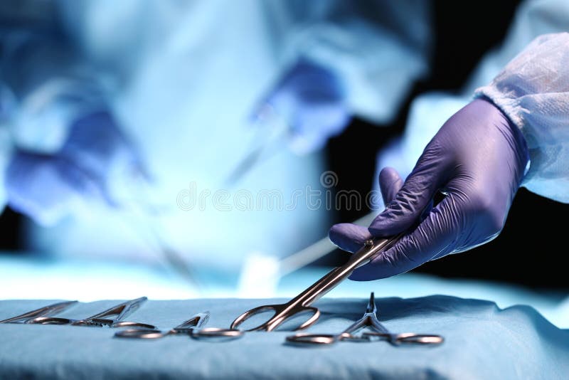 Nurse hand taking surgical instrument for group of surgeons at background operating patient in surgical theatre. Steel medical instruments ready to be used. Surgery and emergency concept. Nurse hand taking surgical instrument for group of surgeons at background operating patient in surgical theatre. Steel medical instruments ready to be used. Surgery and emergency concept