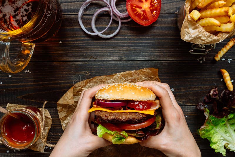 Χέρια που κρατούν τα φρέσκα εύγευστα burgers με τις τηγανιτές πατάτες, τη σάλτσα και την μπύρα στην ξύλινη άποψη επιτραπέζιων κορ στοκ εικόνες Χέρια που κρατούν τα φρέσκα εύγευστα burgers με τις τηγανιτές πατάτες, τη σάλτσα και την μπύρα στην ξύλινη άποψη επιτραπέζιων κορ στοκ εικόνες