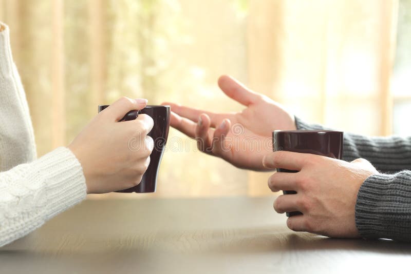 Back light profile of hands of 2 friends or couple talking holding coffee cups sitting in a table at home with a window in the background. Back light profile of hands of 2 friends or couple talking holding coffee cups sitting in a table at home with a window in the background