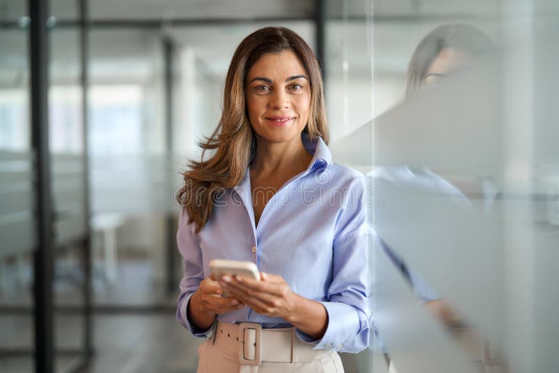 Smiling middle aged woman business investor using mobile apps on cellular phone standing in office. Mature businesswoman executive holding cellphone working on smartphone looking at camera. Portrait. Smiling middle aged woman business investor using mobile apps on cellular phone standing in office. Mature businesswoman executive holding cellphone working on smartphone looking at camera. Portrait.