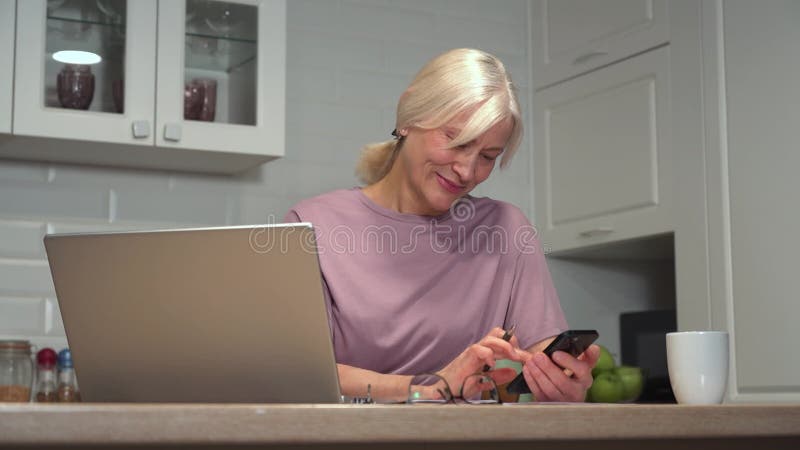 Χαμογελαστή γυναίκα που πληκτρολογεί στο smartphone στη μοντέρνα κουζίνα του σπιτιού απόθεμα βίντεο