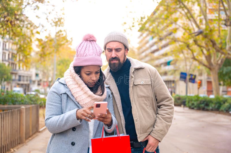 Cute multiethnic couple of man and woman standing holding shopping bags, browsing smartphone apps shopping online. Cute multiethnic couple of man and woman standing holding shopping bags, browsing smartphone apps shopping online