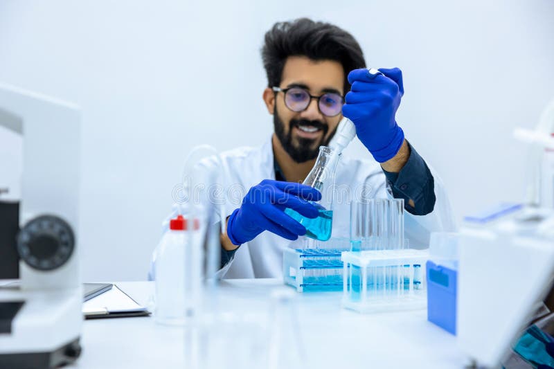 χαμογελαστός άνδρας χημικός με προστατευτικά γάντια που κάθεται στο τραπέζι και κρατά φιάλη ιατρικού δείγματος στοκ εικόνα με δικαίωμα ελεύθερης χρήσης