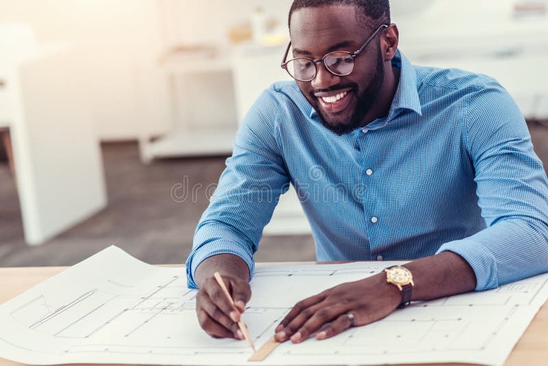 Love my job. Happy African American guy smiling cheerfully while focusing his attention a piece of paper and drawing with a pencil while working on a project alone. Love my job. Happy African American guy smiling cheerfully while focusing his attention a piece of paper and drawing with a pencil while working on a project alone.