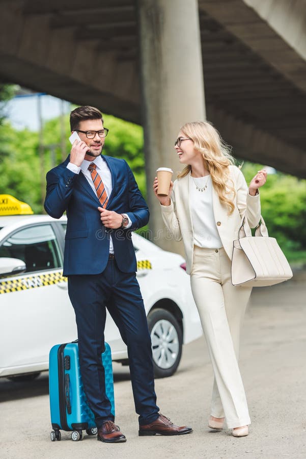 smiling blonde girl holding paper cup and looking at handsome man talking by smartphone while standing with suitcase near taxi. smiling blonde girl holding paper cup and looking at handsome man talking by smartphone while standing with suitcase near taxi