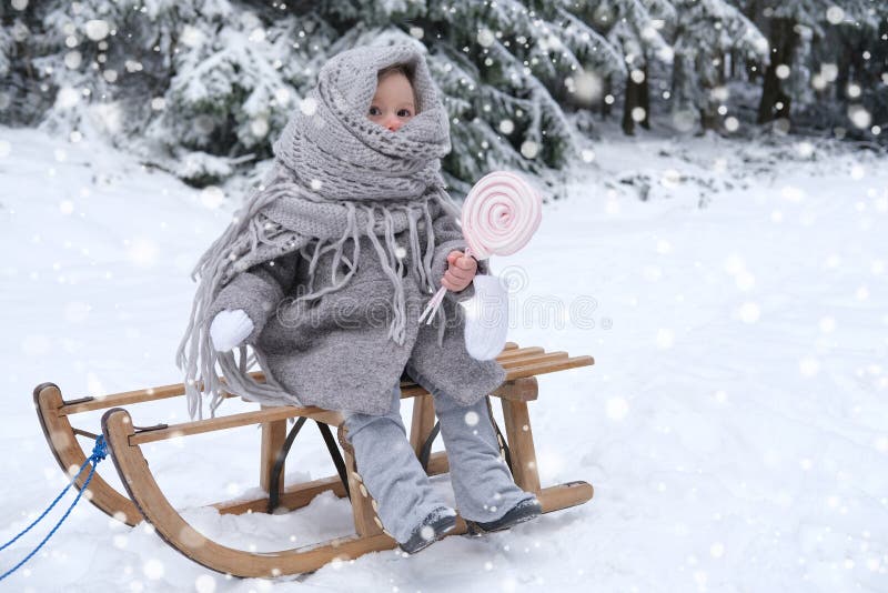 Cute child, girl 2 years old in warm winter retro clothes sits on a wooden sled in the winter forest and eats candy, beautiful landscape, snow-covered fluffy fir trees, walks in the white forest. Cute child, girl 2 years old in warm winter retro clothes sits on a wooden sled in the winter forest and eats candy, beautiful landscape, snow-covered fluffy fir trees, walks in the white forest