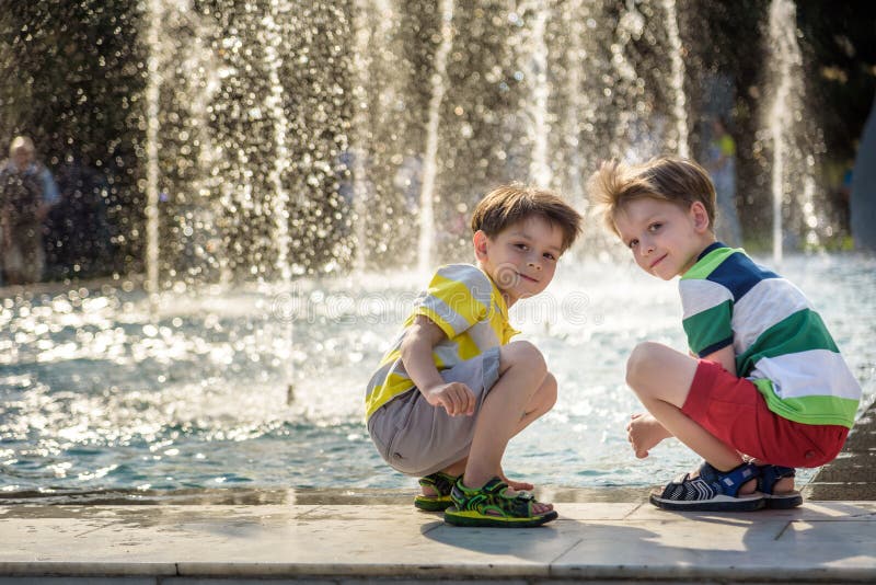 Χαριτωμένο παιδί και μεγαλύτερα αδέρφια, που παίζουν σε ένα συντριβάνι με νερό να τριγυρίζει, καλοκαιρινή ώρα στοκ εικόνες