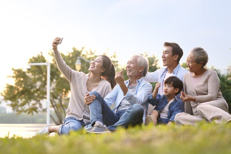 Three generation happy asian family sitting on grass taking a selfie using mobile phone outdoors in park. Three generation happy asian family sitting on grass taking a selfie using mobile phone outdoors in park