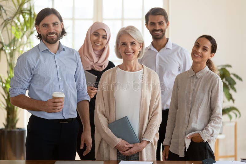 Portrait of pleasant middle aged female boss standing with smiling younger mixed race colleagues. Happy successful international team of professionals posing for photo at office, looking at camera. Portrait of pleasant middle aged female boss standing with smiling younger mixed race colleagues. Happy successful international team of professionals posing for photo at office, looking at camera.