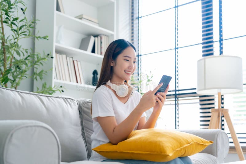 Happy young Asian woman relax sitting on couch using cell phone, smiling lady laughing holding smartphone, looking at cellphone enjoying doing online ecommerce shopping in mobile apps. Happy young Asian woman relax sitting on couch using cell phone, smiling lady laughing holding smartphone, looking at cellphone enjoying doing online ecommerce shopping in mobile apps