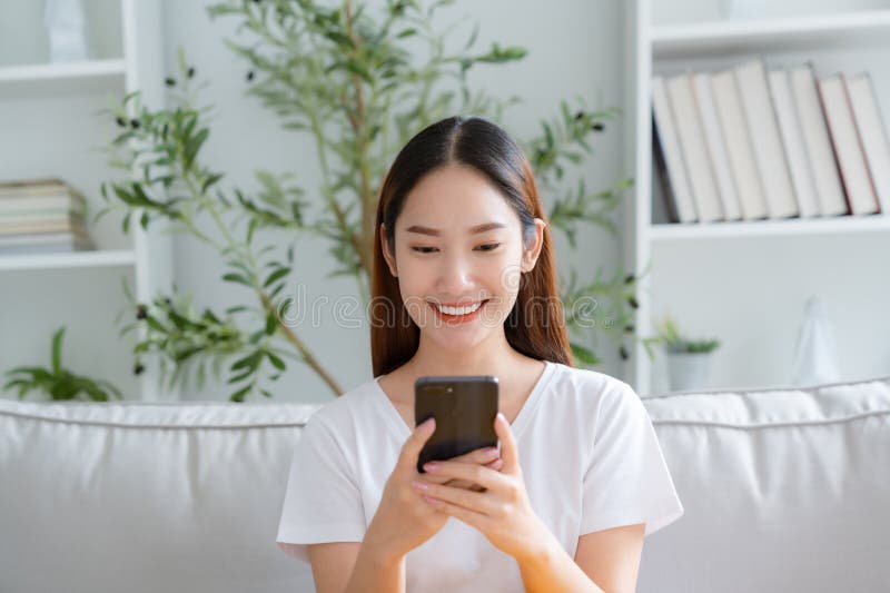 Happy young Asian woman relax sitting on couch using cell phone, smiling lady laughing holding smartphone, looking at cellphone enjoying doing online ecommerce shopping in mobile apps. Happy young Asian woman relax sitting on couch using cell phone, smiling lady laughing holding smartphone, looking at cellphone enjoying doing online ecommerce shopping in mobile apps