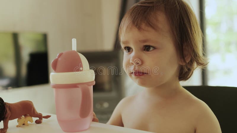 Χαρούμενο νήπιο πόσιμο νερό από ροζ κούπα ή μπουκάλι με ένα καλαμάκι περιτριγυρισμένο από παιχνίδια που κάθονται σε ένα τραπέζι σε απόθεμα βίντεο