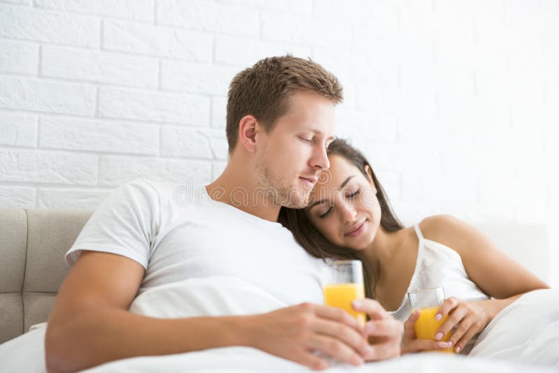 Happy young couple handsome men and beautiful women lying in bed in bright bedroom in the morning drinking orange juice family idyll. Happy young couple handsome men and beautiful women lying in bed in bright bedroom in the morning drinking orange juice family idyll.