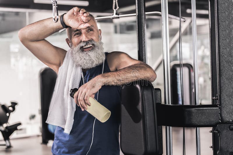 Tired and happy. Strong muscular old bearded man is listening to music through earphones with smile and holding bottle of water while wiping his brow with hand in modern gym. Tired and happy. Strong muscular old bearded man is listening to music through earphones with smile and holding bottle of water while wiping his brow with hand in modern gym