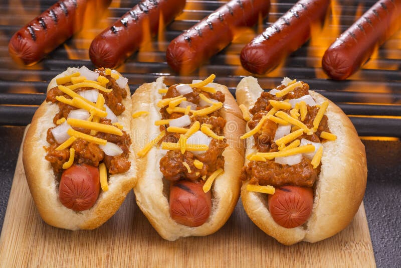 Three chili hot dogs with cheese and onions in buns Grilled with hot dogs on a hot flaming grill in the background. Three chili hot dogs with cheese and onions in buns Grilled with hot dogs on a hot flaming grill in the background.