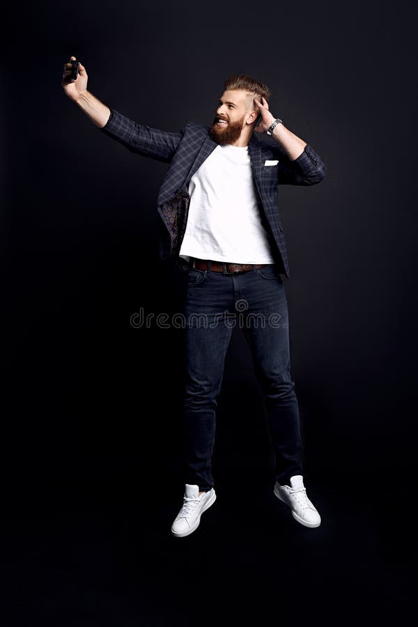 Χρόνος για λήψη της selfie Σε όλο το μήκος ενός όμορφου νεαρού με γενειάδα, με ρούχα της μόδας, που παίρνει selfie ενώ πηδάει ενά στοκ φωτογραφία με δικαίωμα ελεύθερης χρήσης