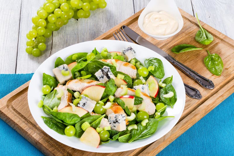 Grilled chicken breast, grapes, celery, spinach, cheese and apple salad on a white dish on a cutting board with fork and knife, view from above, close-up. Grilled chicken breast, grapes, celery, spinach, cheese and apple salad on a white dish on a cutting board with fork and knife, view from above, close-up