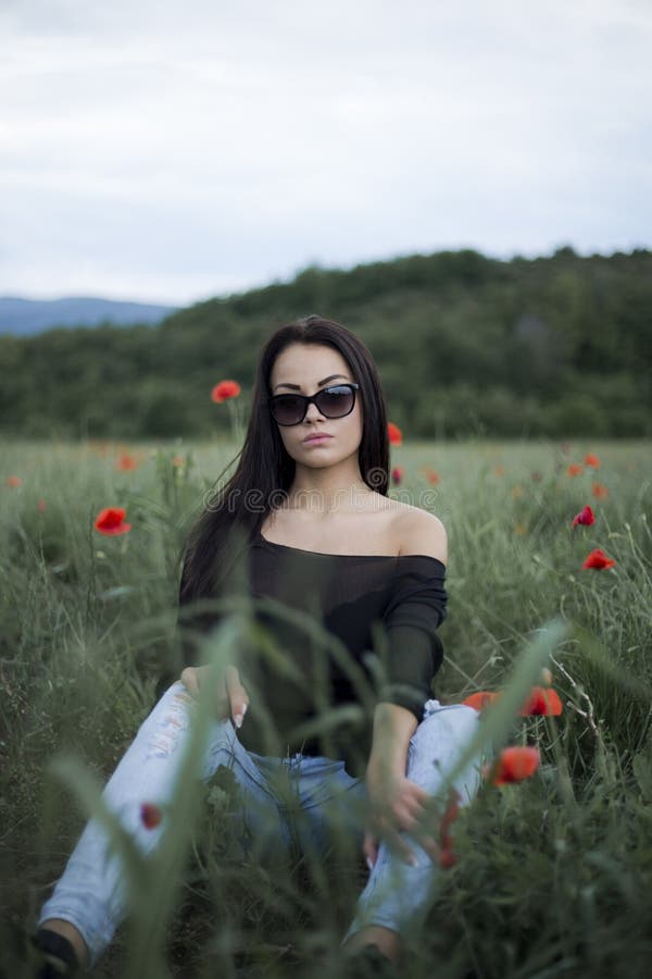 Όμορφη γυναίκα brunette στα γυαλιά ηλίου και τα μοντέρνα ενδύματα που κάθεται μεταξύ στις κόκκινες παπαρούνες στοκ εικόνες Όμορφη γυναίκα brunette στα γυαλιά ηλίου και τα μοντέρνα ενδύματα που κάθεται μεταξύ στις κόκκινες παπαρούνες στοκ εικόνες
