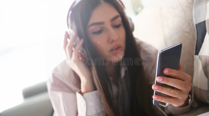 Young beautiful woman headphones holds smartphone in hand using at home costs about sofa. Verifies internal information its financial advertising freedom makes money level transfer contractor concept. Young beautiful woman headphones holds smartphone in hand using at home costs about sofa. Verifies internal information its financial advertising freedom makes money level transfer contractor concept