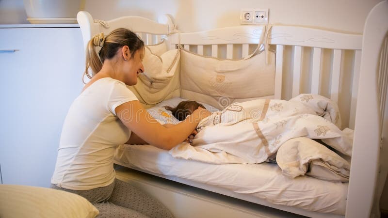 Όμορφη νέα μητέρα στις πυτζάμες που χαϊδεύει το χαριτωμένο ύπνο γιων μικρών παιδιών της στο παχνί στο βρεφικό σταθμό στοκ φωτογραφία με δικαίωμα ελεύθερης χρήσης Όμορφη νέα μητέρα στις πυτζάμες που χαϊδεύει το χαριτωμένο ύπνο γιων μικρών παιδιών της στο παχνί στο βρεφικό σταθμό στοκ φωτογραφία με δικαίωμα ελεύθερης χρήσης
