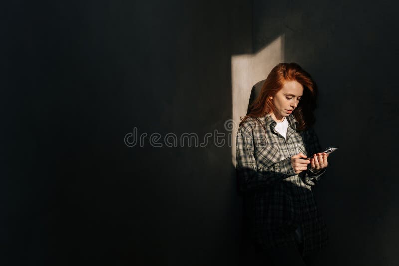 Beautiful young woman holding using mobile phone at shadow background in dark living room, looking to smartphone screen, enjoying doing online ecommerce shopping in mobile apps indoors. Beautiful young woman holding using mobile phone at shadow background in dark living room, looking to smartphone screen, enjoying doing online ecommerce shopping in mobile apps indoors.