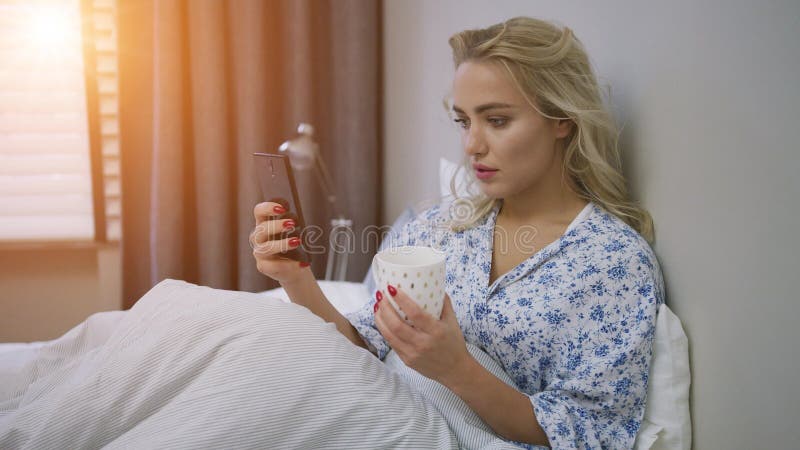 όμορφη νεαρή γυναίκα που περιηγείται το smartphone και κρατά μια κούπα ζεστό ρόφημα στο κρεβάτι απόθεμα βίντεο
