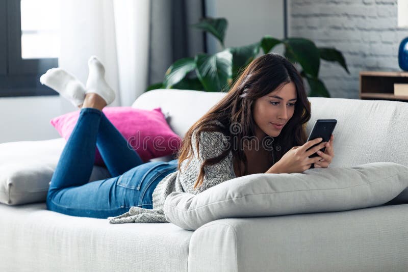 Όμορφη νεαρή γυναίκα στέλνει μηνύματα με το κινητό της ενώ ξαπλώνει στον καναπέ στο σπίτι στοκ φωτογραφία με δικαίωμα ελεύθερης χρήσης Όμορφη νεαρή γυναίκα στέλνει μηνύματα με το κινητό της ενώ ξαπλώνει στον καναπέ στο σπίτι στοκ φωτογραφία με δικαίωμα ελεύθερης χρήσης