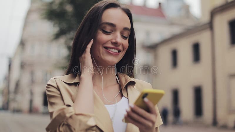 Όμορφη νεαρή κοπέλα με ακουστικά απολαμβάνει να ακούει μουσική χρησιμοποιώντας το smartphone της και χαμογελώντας περπατώντας στη