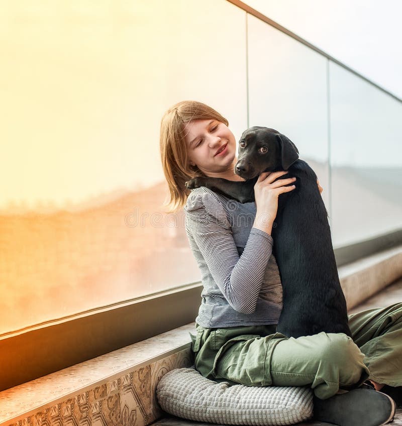 A pretty teenage girl hugs her black dog on the terrace of a solitaire in the city. A pretty teenage girl hugs her black dog on the terrace of a solitaire in the city