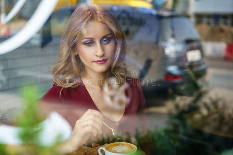 Όμορφη συνεδρίαση γυναικών από το παράθυρο σε έναν καφέ κατανάλωσης καφέδων στοκ φωτογραφία Όμορφη συνεδρίαση γυναικών από το παράθυρο σε έναν καφέ κατανάλωσης καφέδων στοκ φωτογραφία
