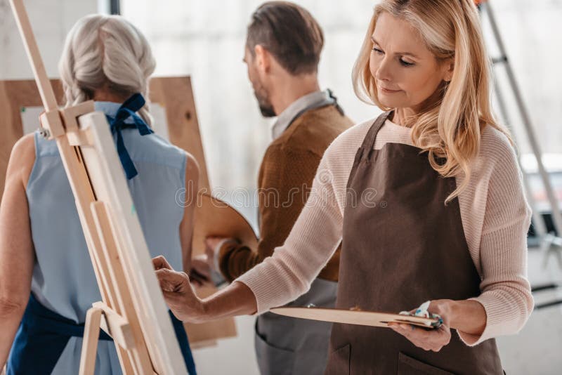 όμορφη ώριμη παλέτα εκμετάλλευσης γυναικών και ζωγραφική easel κατά τη διάρκεια της κατηγορίας τέχνης στοκ φωτογραφία όμορφη ώριμη παλέτα εκμετάλλευσης γυναικών και ζωγραφική easel κατά τη διάρκεια της κατηγορίας τέχνης στοκ φωτογραφία