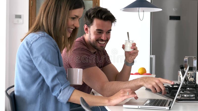 Όμορφο καλό νέο ζεύγος χρησιμοποιώντας το lap-top τους και έχοντας το πρόγευμα στην κουζίνα στο σπίτι φιλμ μικρού μήκους