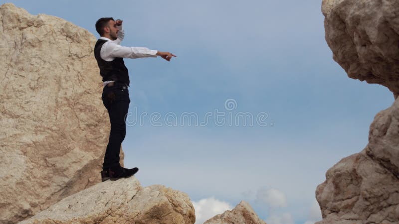 Όμορφο μοντέρνο άτομο στο φανταχτερό ύφασμα που στέκεται στους βράχους και που μια καταπληκτική άποψη παρουσιάζοντας κάτι στους α