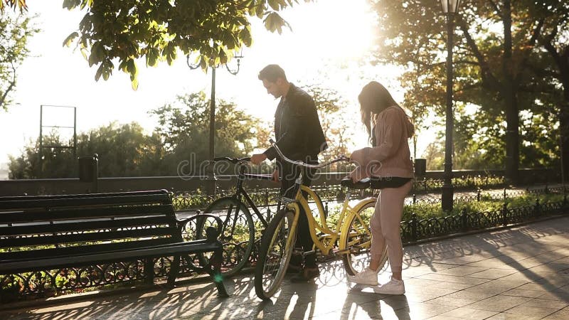 Όμορφο νέο ζεύγος που οδηγά τα ποδήλατά τους στο κενό πάρκο πόλεων Η στάση και κάθεται στον πάγκο και το αγκάλιασμα αγάπη ζευγών