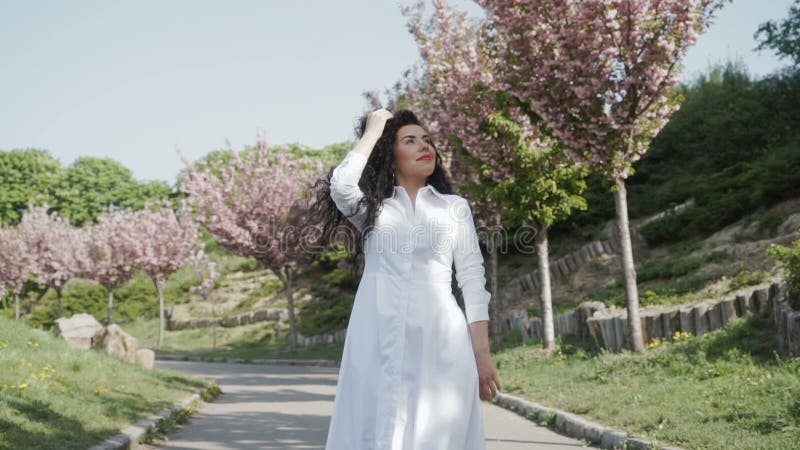 Όμορφο σγουρό brunette στο άσπρο φόρεμα που περπατά στο ανθίζοντας πάρκο