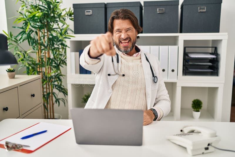 Handsome middle age doctor man working at the clinic pointing displeased and frustrated to the camera, angry and furious with you. Handsome middle age doctor man working at the clinic pointing displeased and frustrated to the camera, angry and furious with you