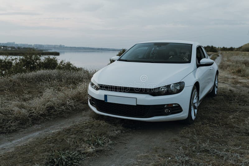 Modern luxury white sedan 2-doors  sport car at the sunset outdoors. Fresh weekend out of city near riverside at countryside. Dark tone photo. Modern luxury white sedan 2-doors  sport car at the sunset outdoors. Fresh weekend out of city near riverside at countryside. Dark tone photo