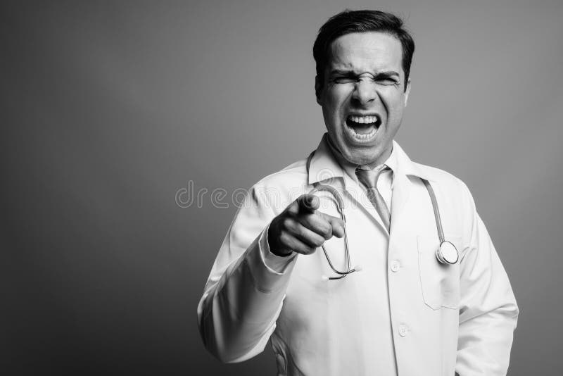 Studio shot of handsome Persian man doctor against gray background in black and white. Studio shot of handsome Persian man doctor against gray background in black and white